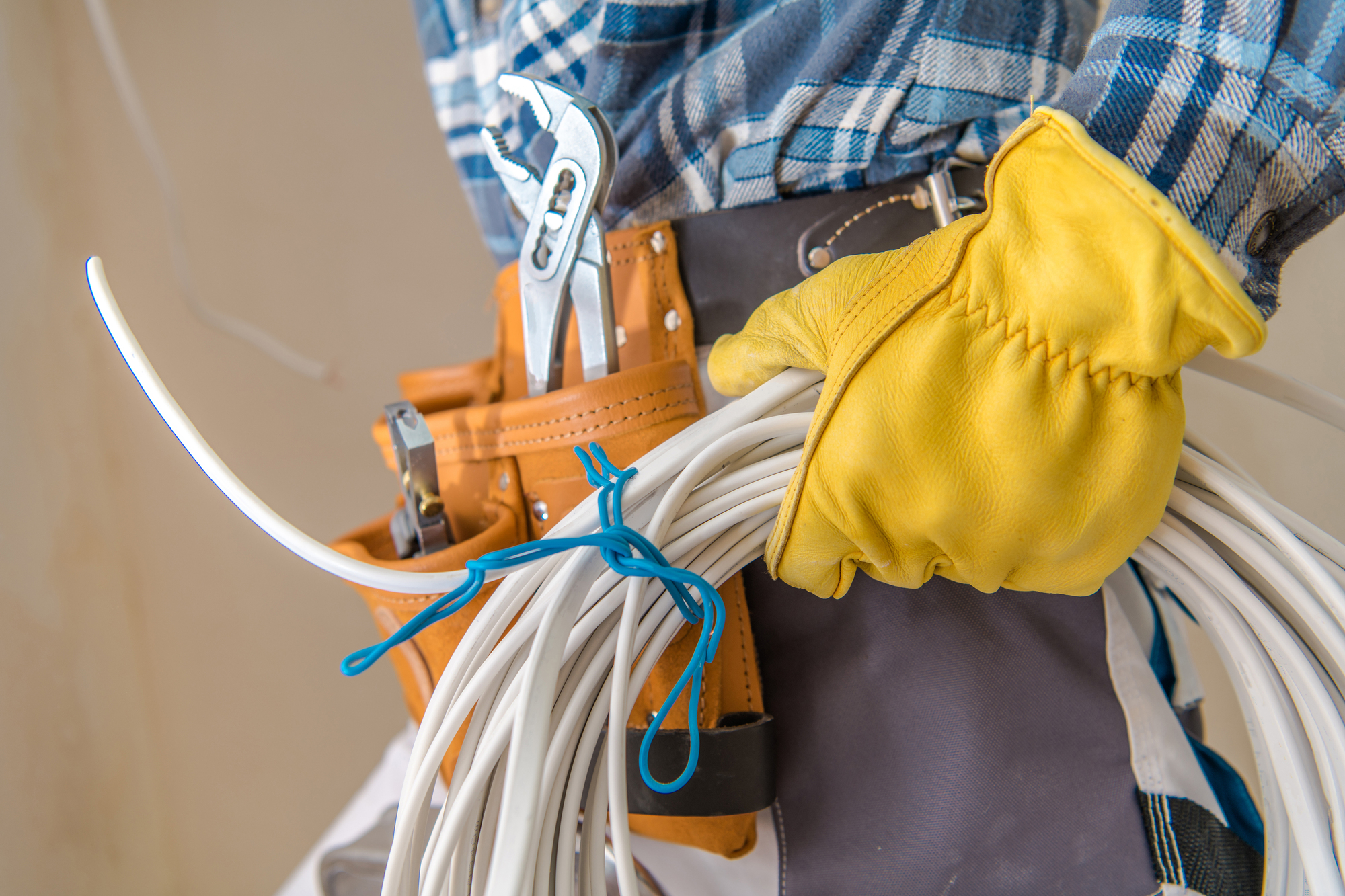 A person wearing yellow gloves holds a bundle of white cables. A tool belt with pliers and tools is visible around their waist.