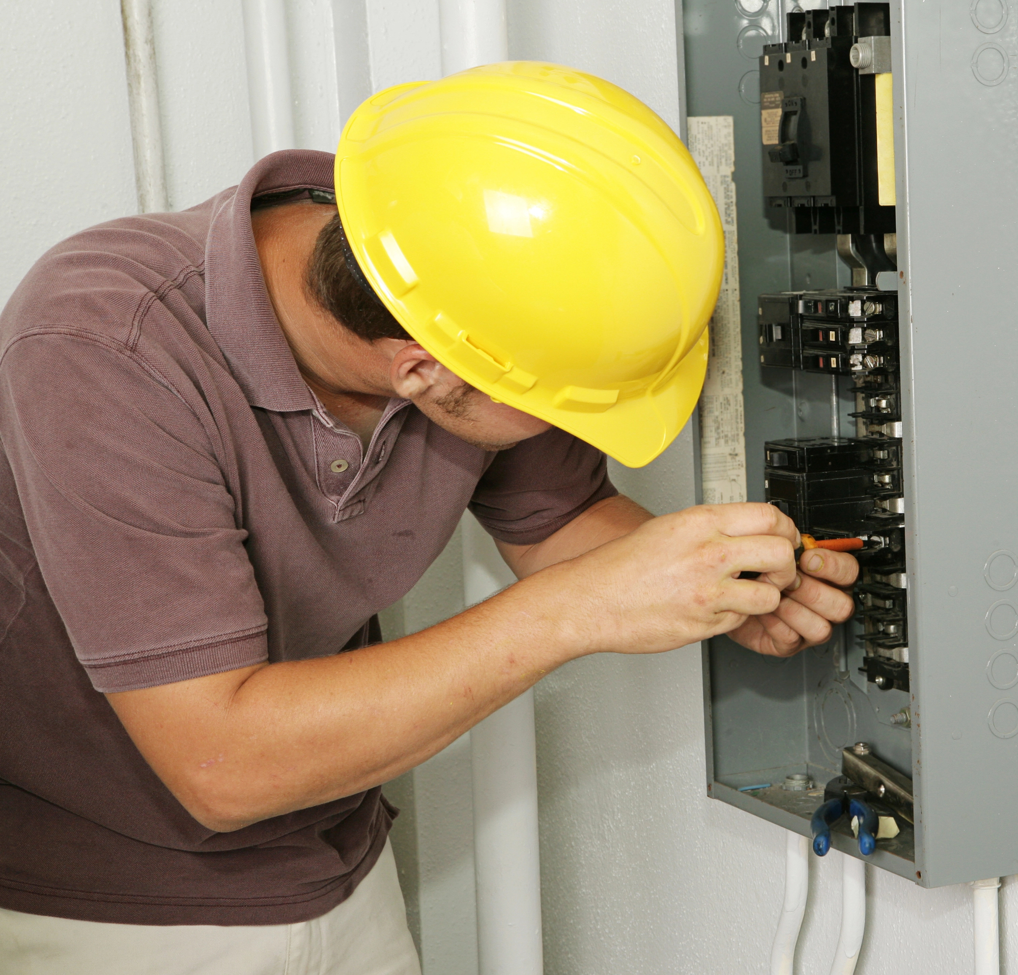 A person wearing a yellow hard hat and a brown shirt is repairing an electrical panel using a screwdriver.