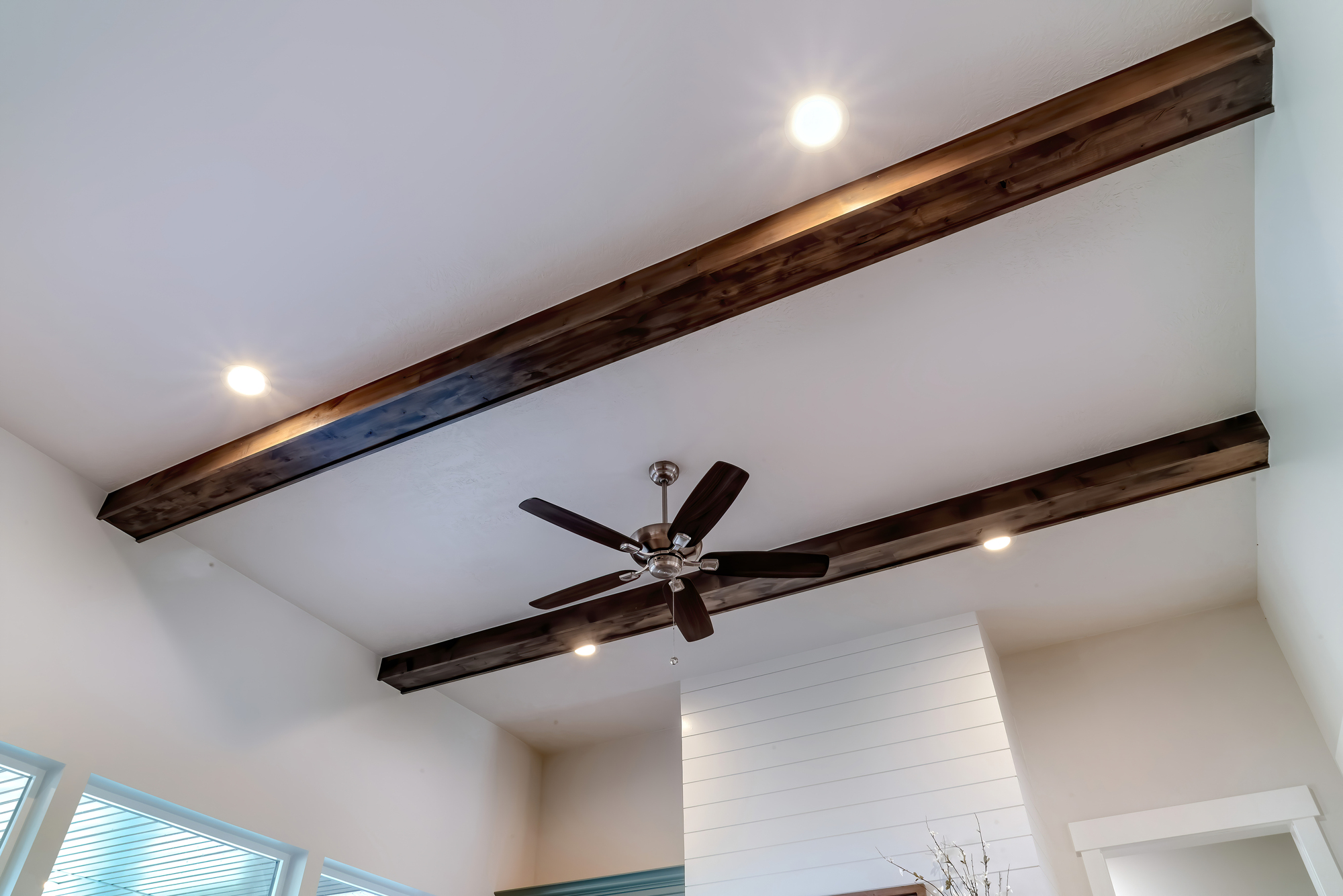 Ceiling with two dark wooden beams, a dark brown ceiling fan, and four round recessed lights in a room with white walls and a window.