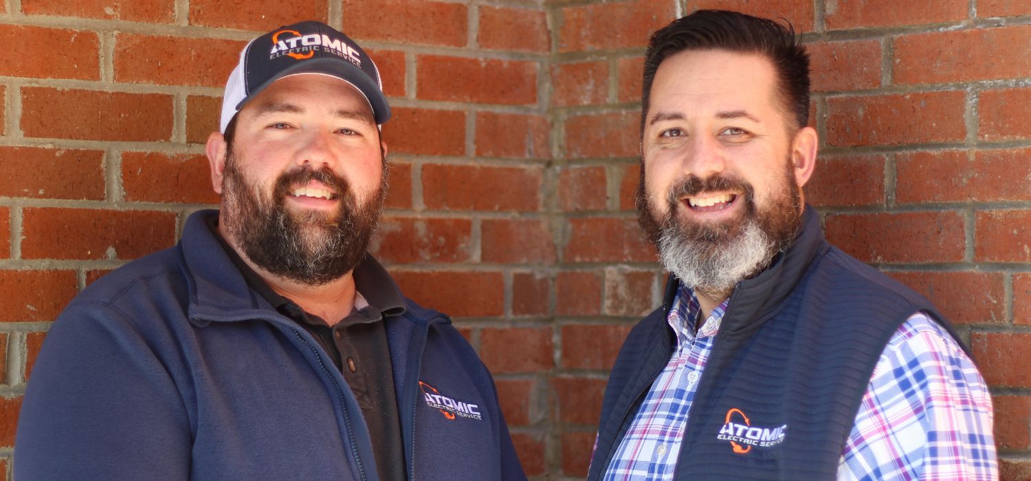 Two owners of Atomic Electric Service Inc. smiling in branded apparel while standing in front of a brick wall.
