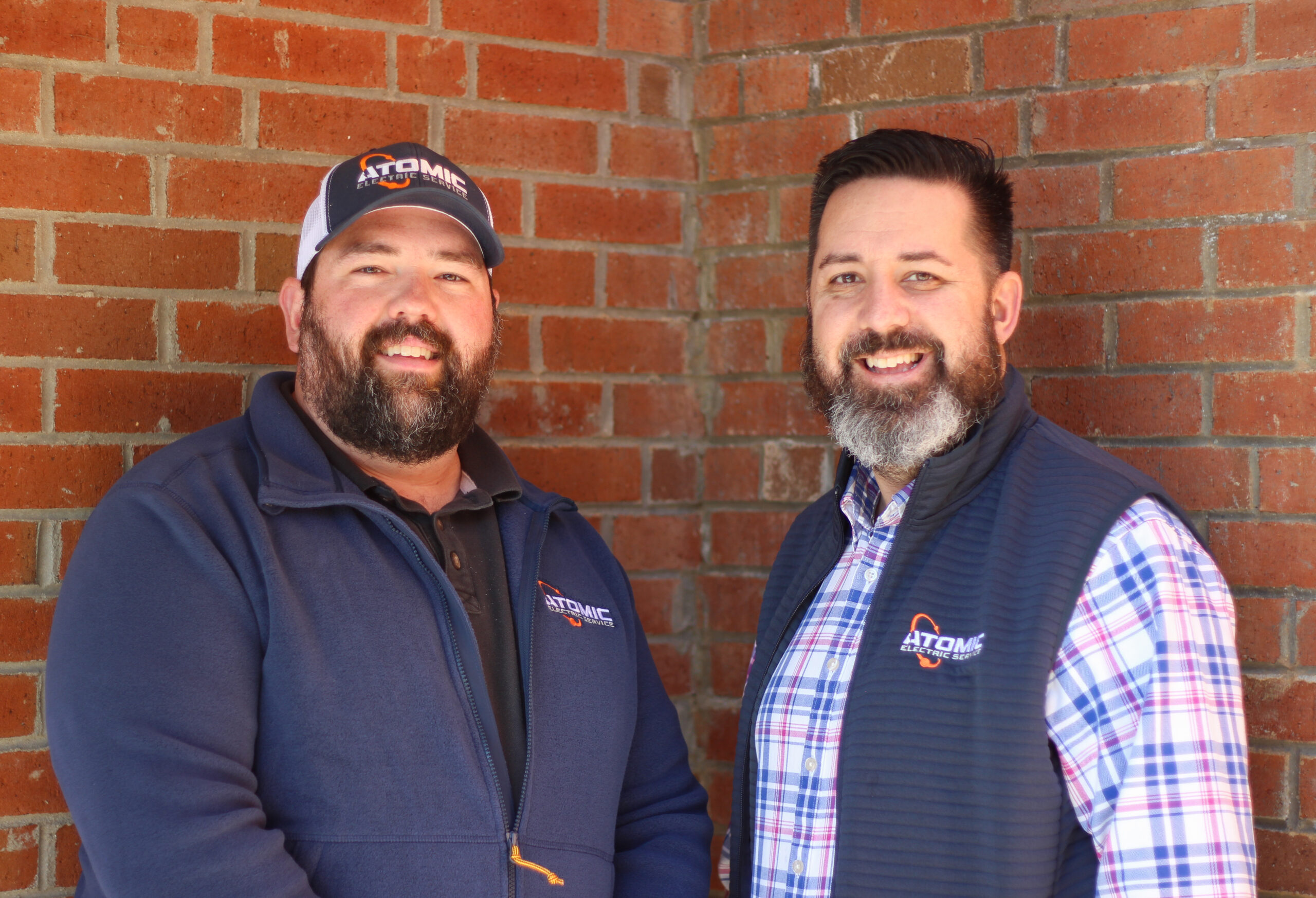 Two men with beards stand in front of a brick wall, both wearing jackets and smiling, capturing the essence of camaraderie that's at the heart of our About Us story.